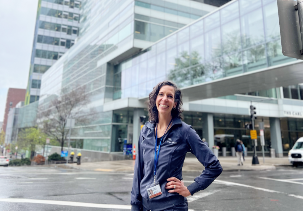 Nurse in grey scrubs stands in front of tall glass buildings
