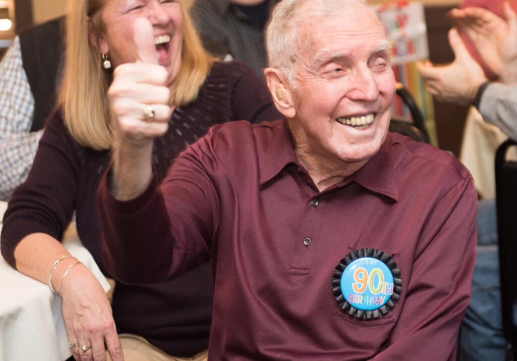 An elderly man celebrating his 90th birthday smiles broadly and gives a thumbs-up while sitting at a table surrounded by friends and family. He is wearing a maroon shirt with a “90th Birthday” badge on his chest. The atmosphere is joyful, with people laughing and clapping in the background.