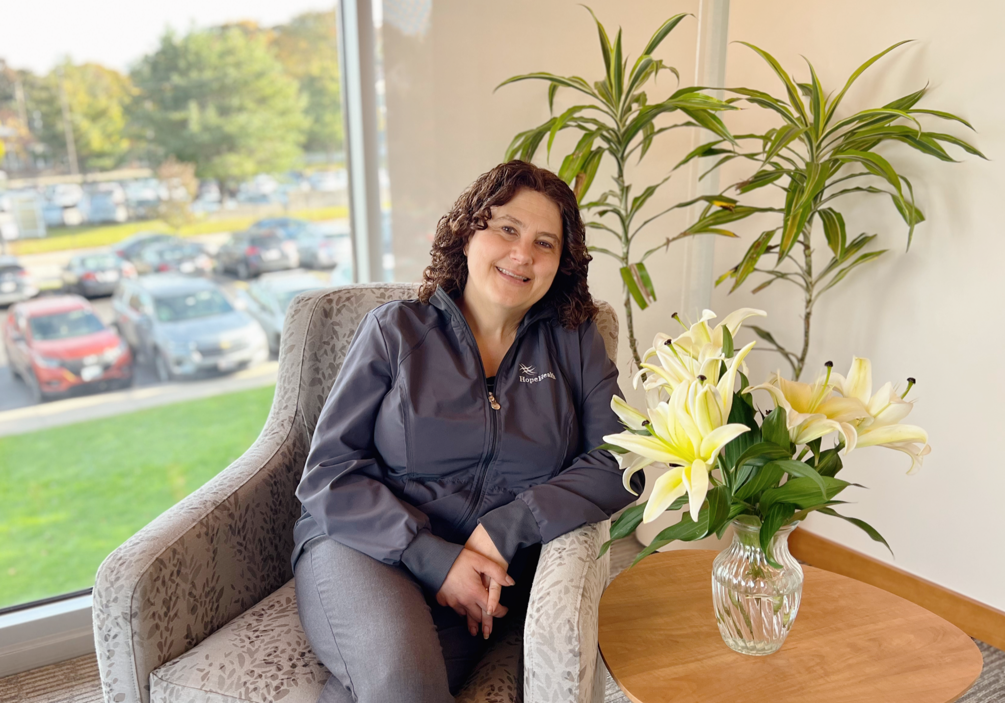 female palliative social worker with brown curly hair sitting in a arm chair next to a vase of white lillies