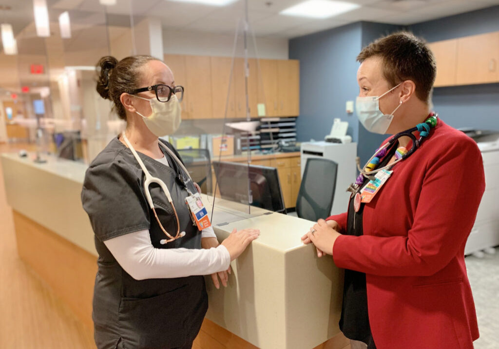 A nurse wearing grey scrubs and a surgical mask chats with a doctor wearing a red blazer and a surgical mask at the Hulitar Hospice Center