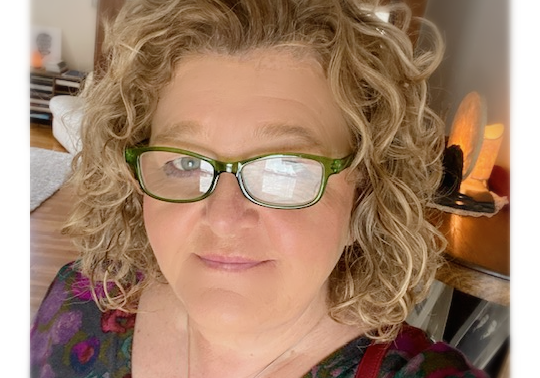 IMG_2206 Blonde woman with curly hair and glasses smiles in a dimly lit room