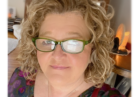 Blonde woman with curly hair and glasses smiles in a dimly lit room