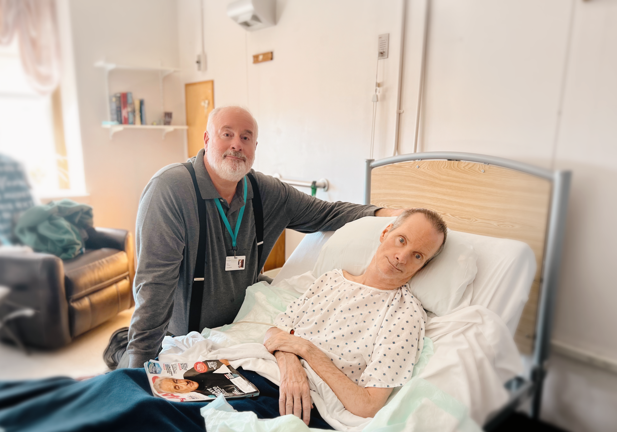 A man in a gray shirt and suspenders compassionately sits by a male hospice patient who is laying in a hospital bed