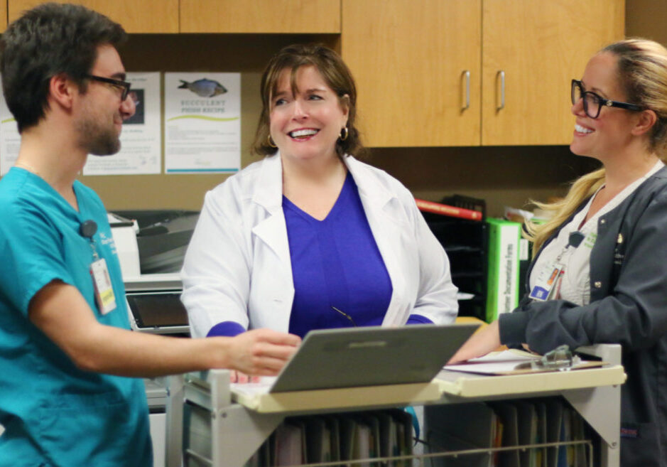 Young male in teal scrubs talking to two female nurses at the nurses station while smiling