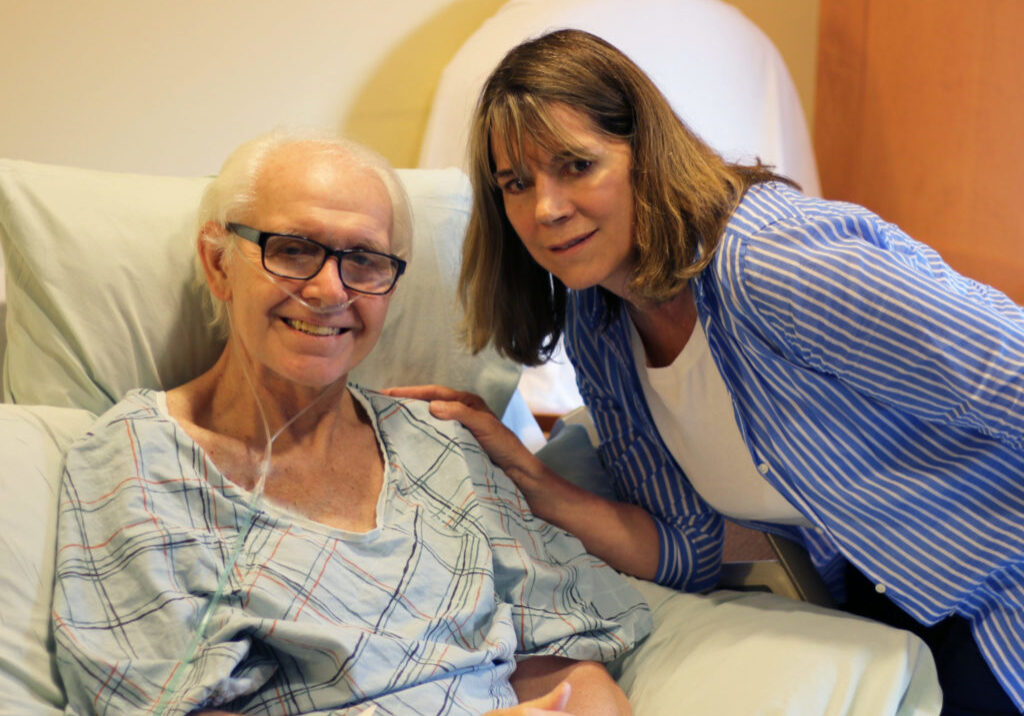 A man on hospice services wearing a hospital gown, in the inpatient unit smiles about his hospice experience, next to his supportive wife who leans close to him.
