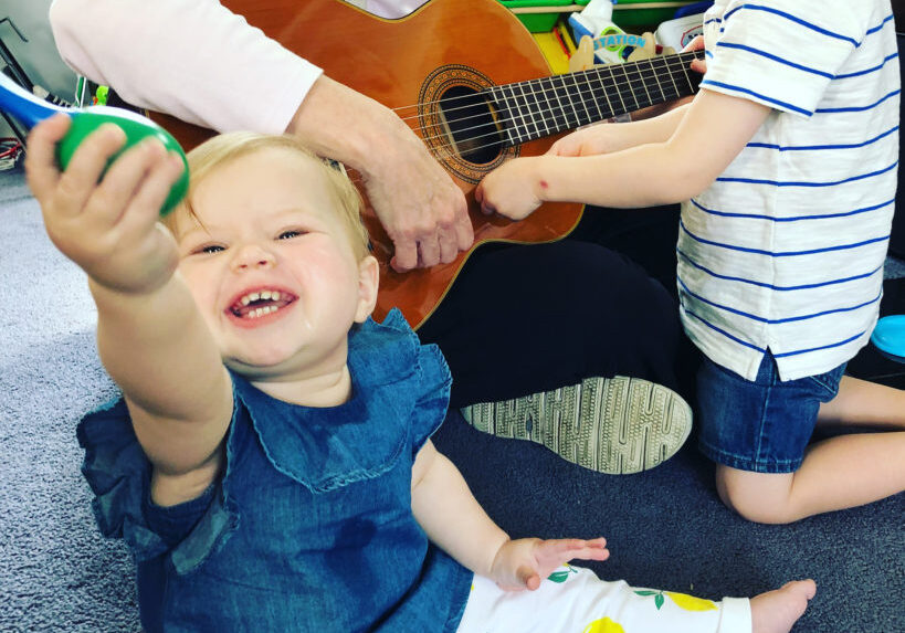 baby smiles and toddler is interested in guitar played by older woman