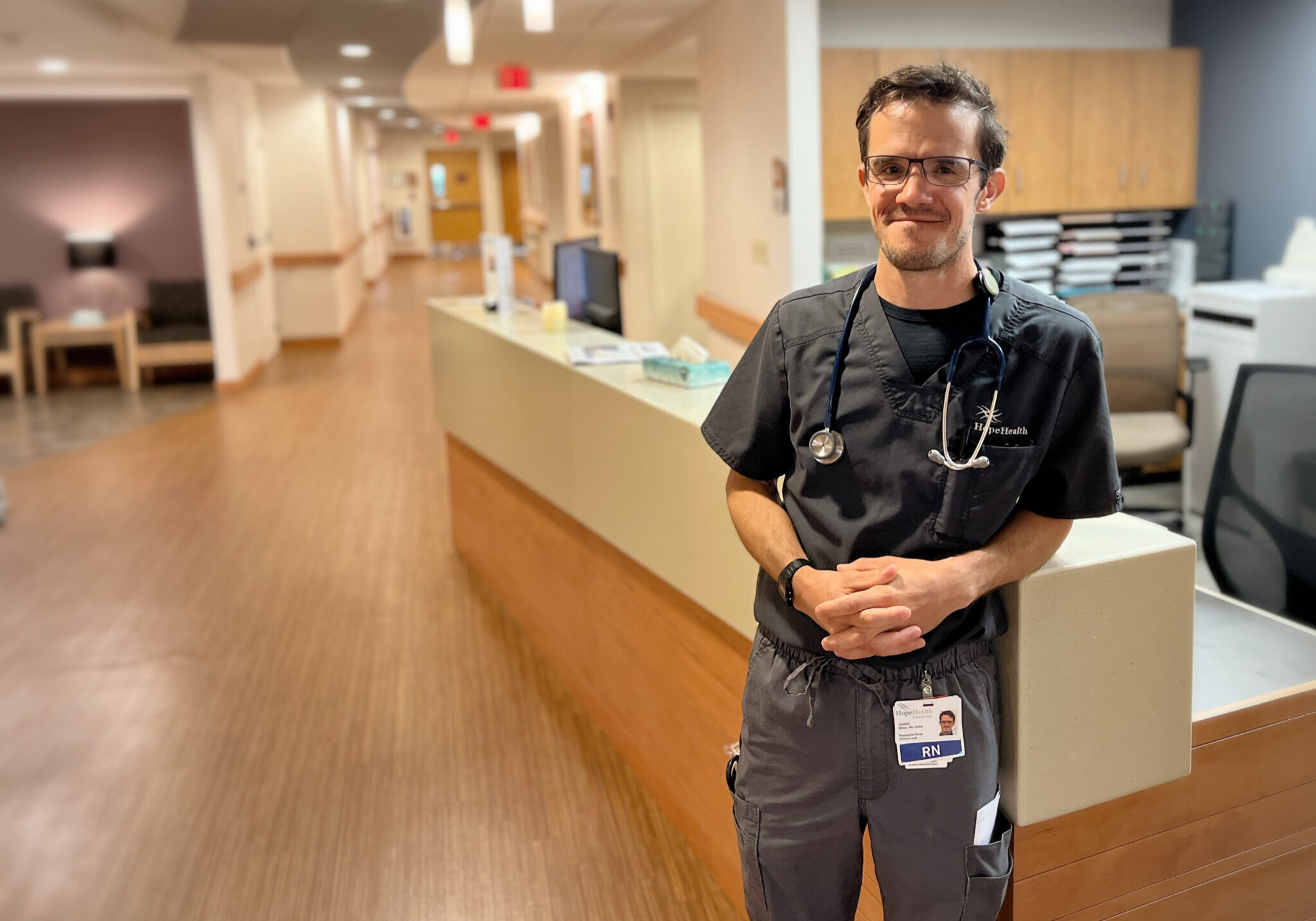 A male nurse stands against a nursing station in a warmly lit hospice center