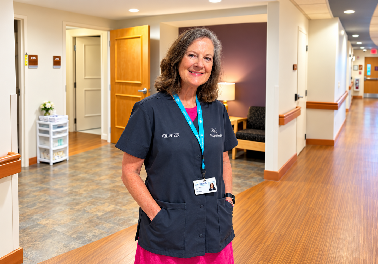 Woman in pink dress and gray volunteer smock smils while standing in front of hospice IPU rooms