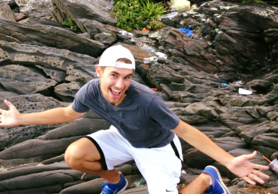 Young man with backwards white cap kneeling on ocean rocks with his hands stretched out