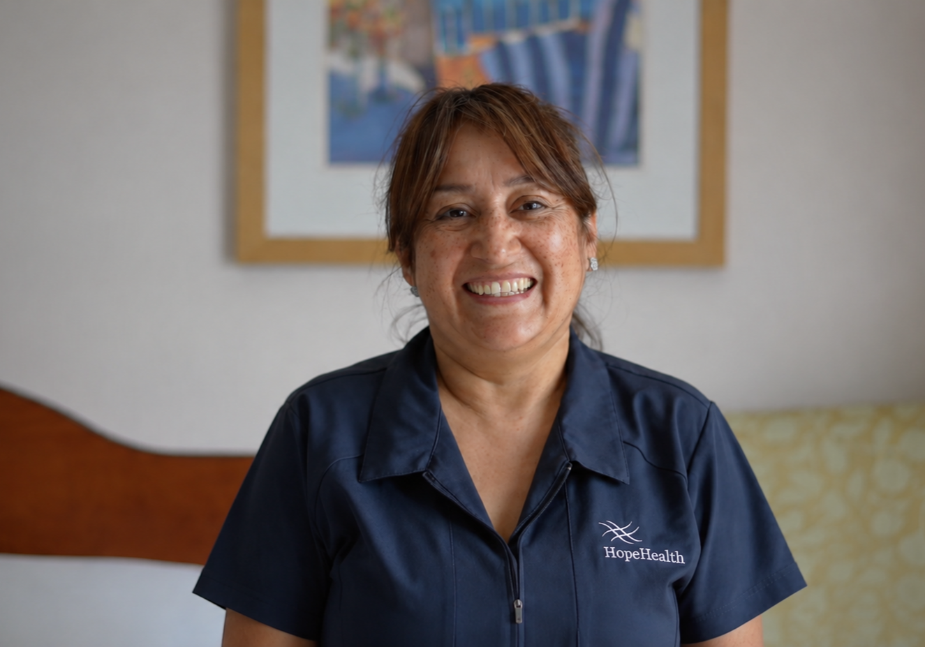 Nora Navas, CNA, stands in a patient room at the HopeHealth Huliar Hospice Center wearing gray scrubs, smiling warmly