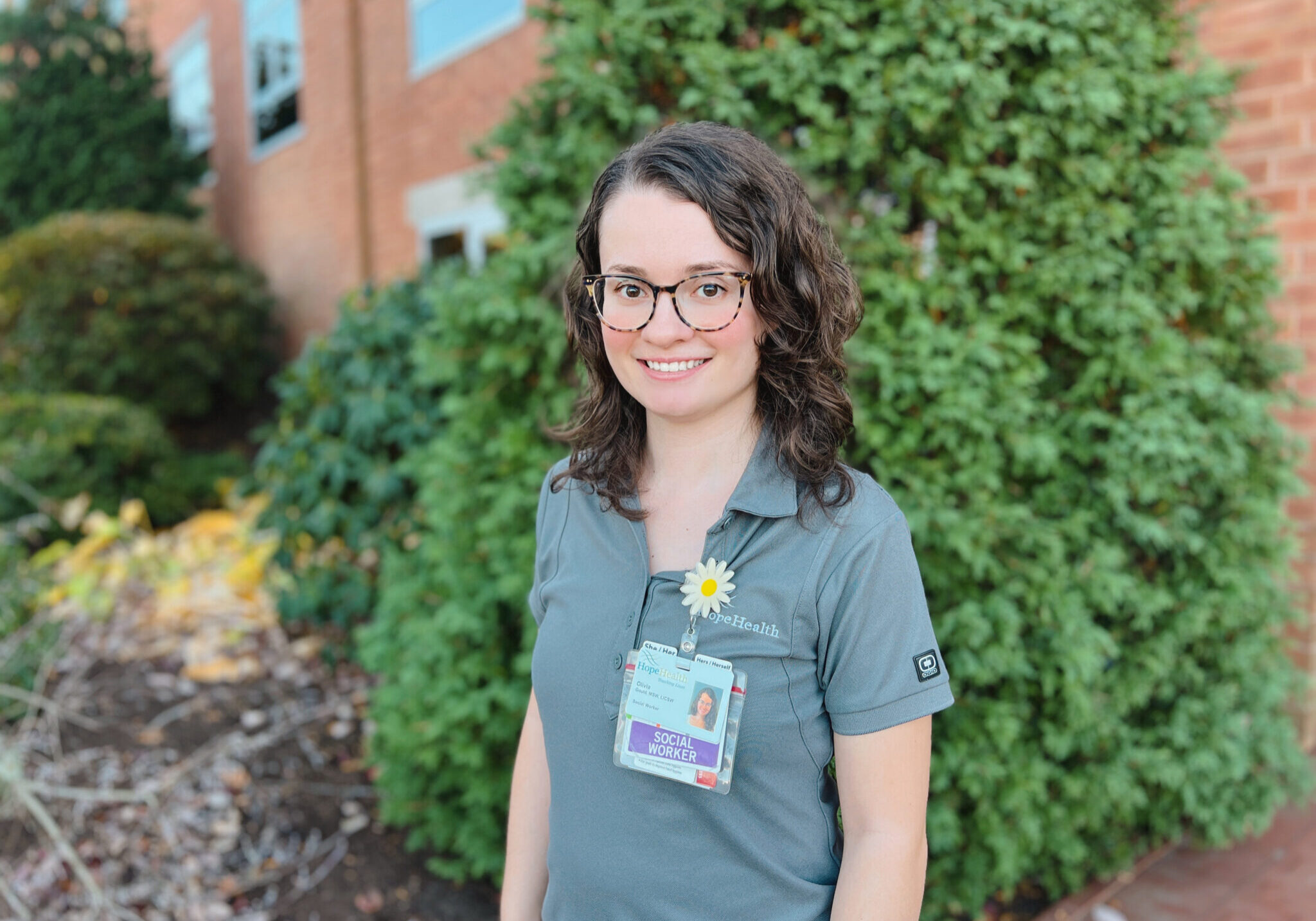Smiling woman with curly brown hair wearing glasses and a gray HopeHealth polo shirt, standing outside in front of greenery. She is wearing a name badge and a daisy flower pin.