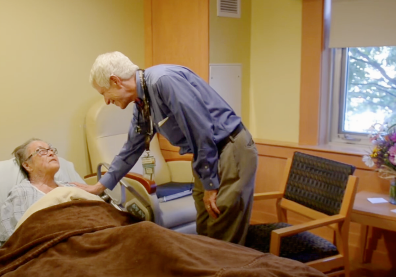 Doctor compassionately leans down to a patient in her bed, reaching out his arm in compassion