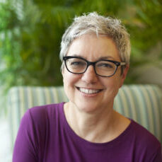 Woman with short gray hair wearing glasses and a deep purple top smiles with greenery in the background