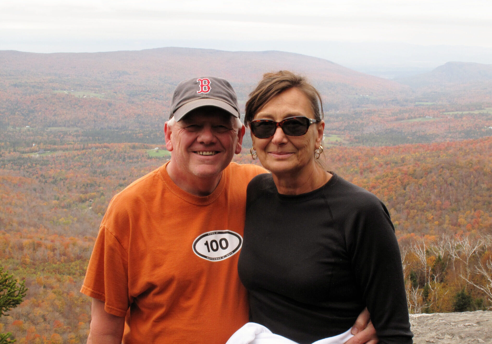 A man and woman embrace in front of a majestic outlook of orange foliage over a mountain range