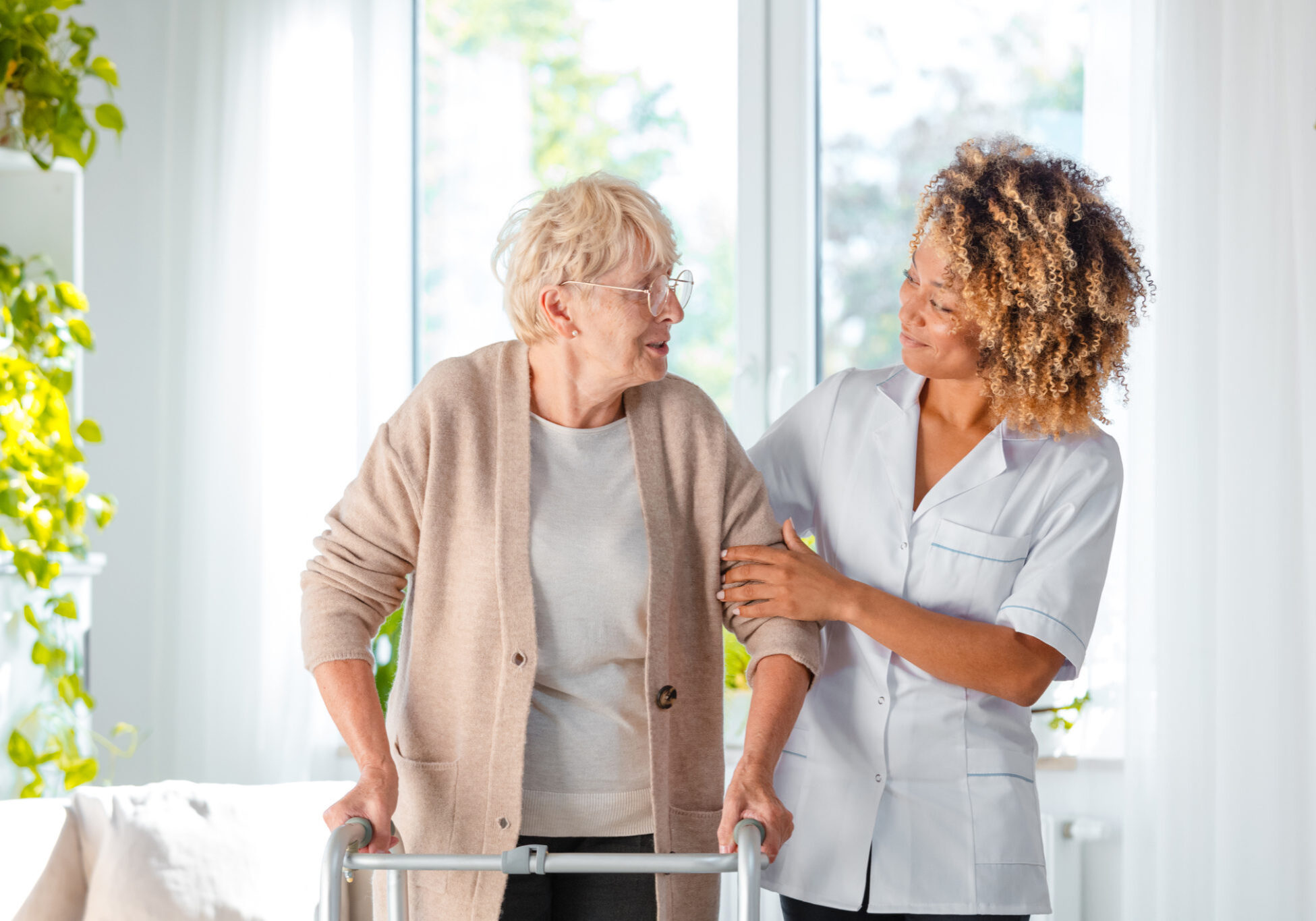 Afro american female home nurse supporting senior woman in using mobility walker at home.