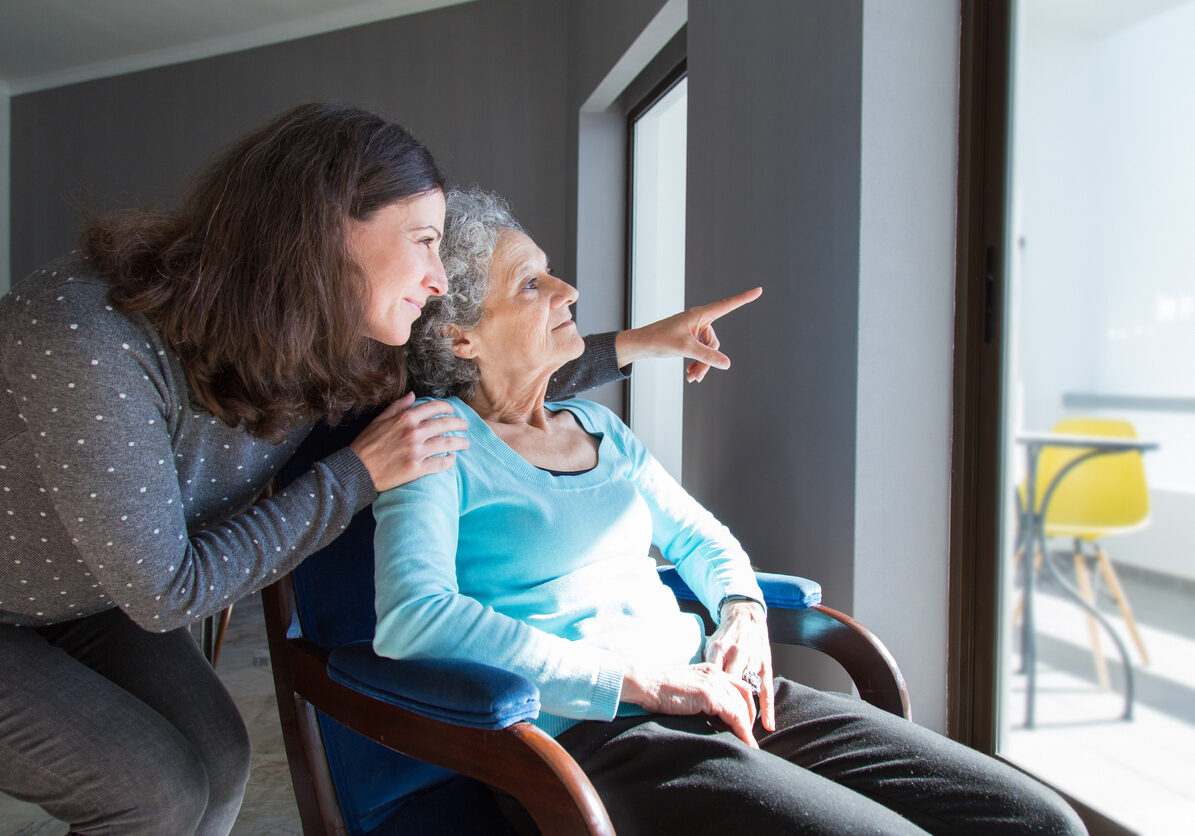 Adult daughter showing to senior mother scene out of window. Young and elderly women looking out window. Assisting concept