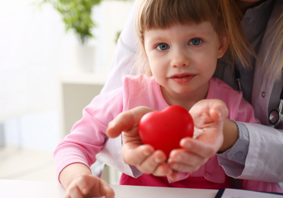 Little baby girl visiting doctor holding in hands red toy heart as life safe symbol