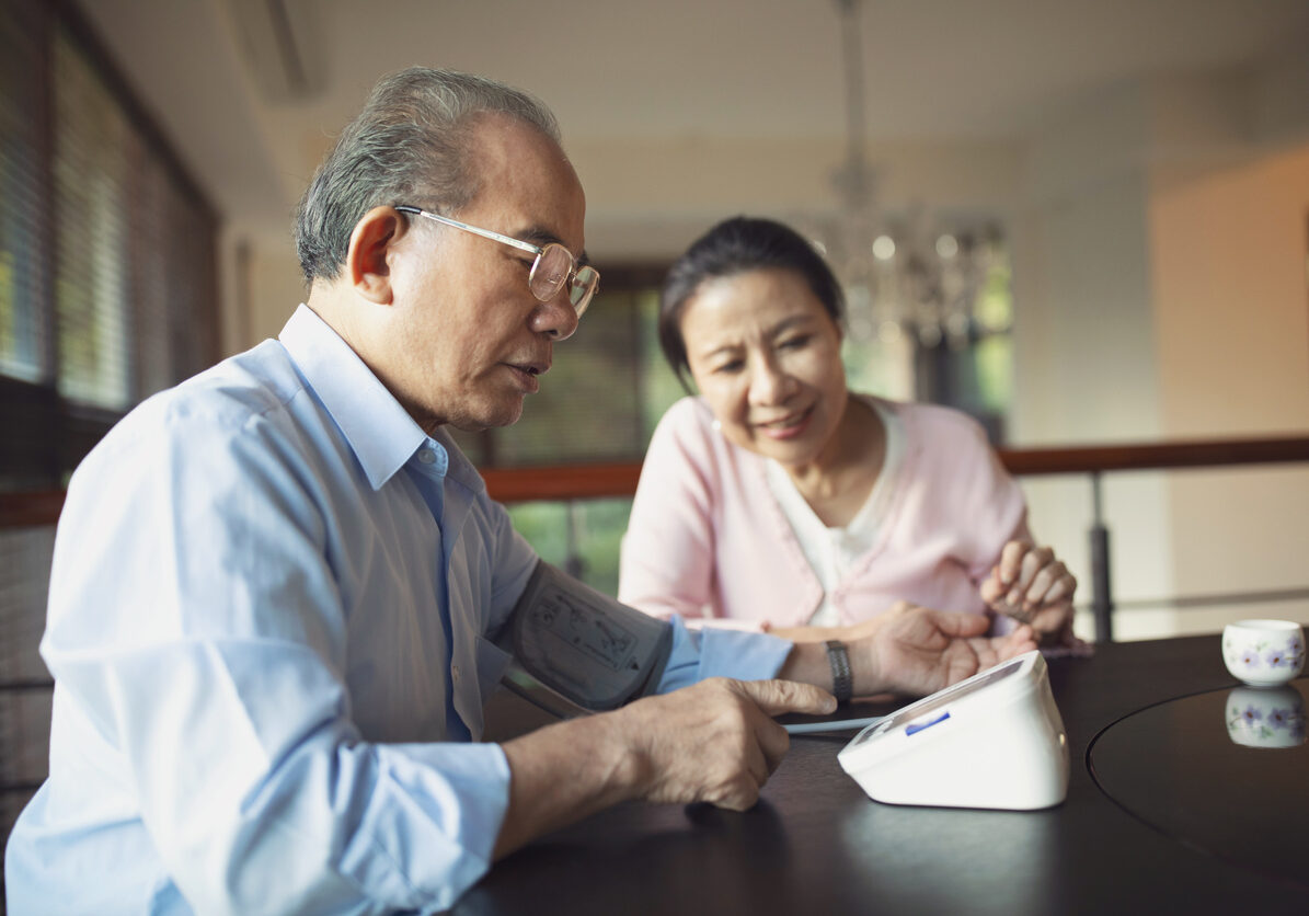 Senior Asian man monitoring his blood pressure at home and his wife sitting next to him.