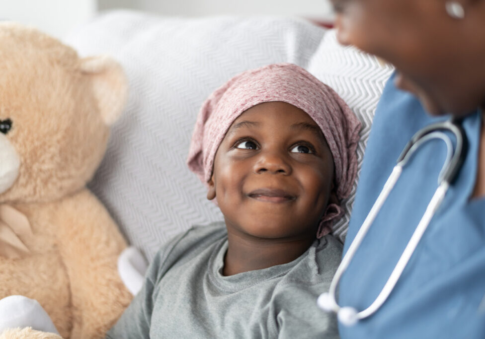 A young boy of African descent is at a medical consultation. He is dressed in casual clothes while wearing a headscarf to hide his hair loss. He sits beside his stuffed animal while listening to his doctor of African descent and smiling up at her.