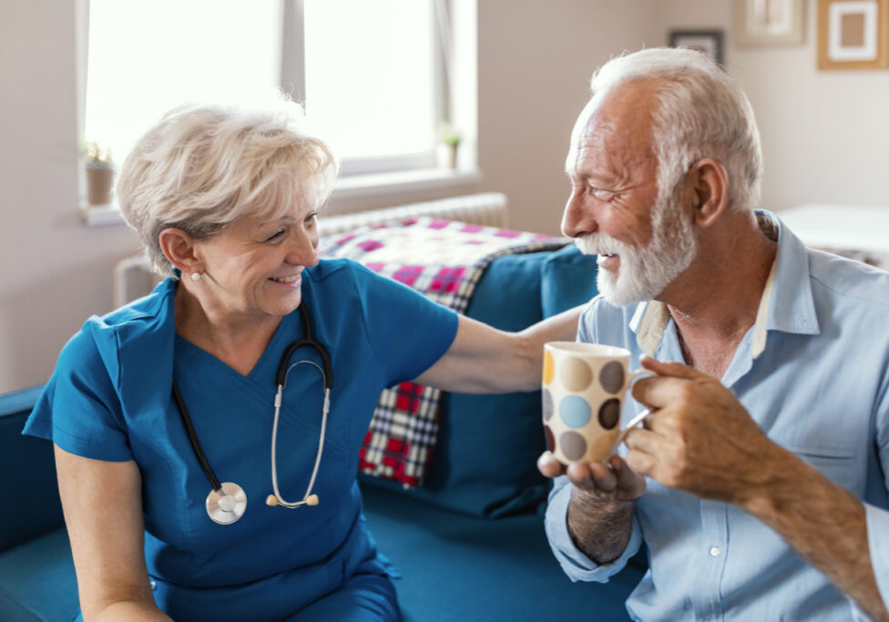 Float hospice nurse smiling at gray-haired male patient drinking from a mug.