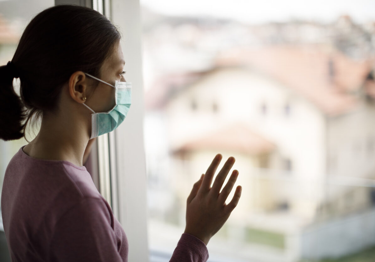 Portrait of sad teenage girl with face protective mask looking through the window at home grieving during coronavirus COVID-19