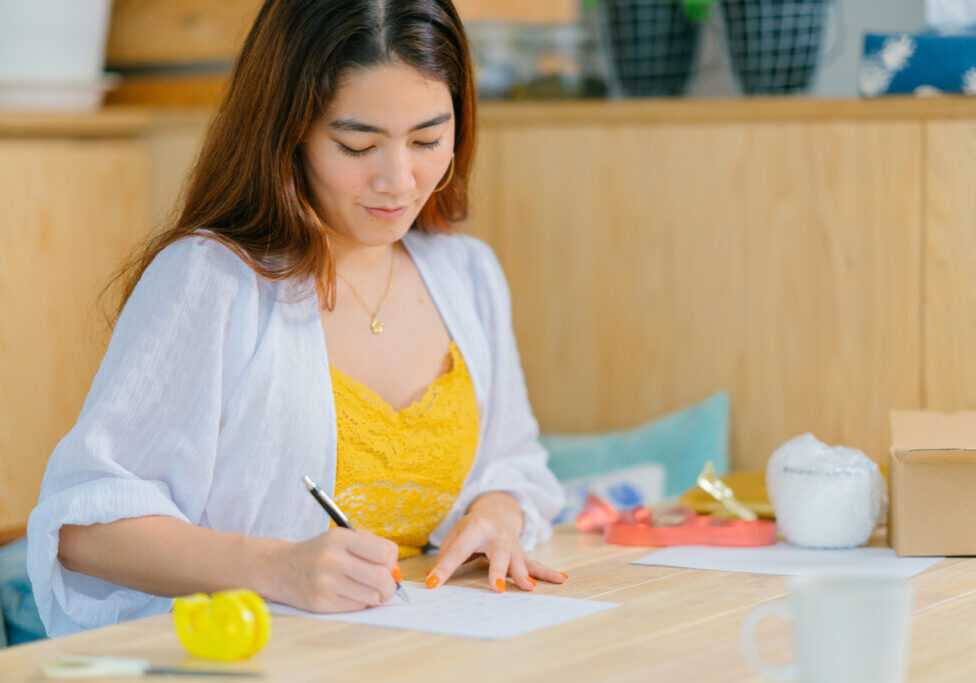 A young woman is writing a letter to put inside a gift box in the living room at home.