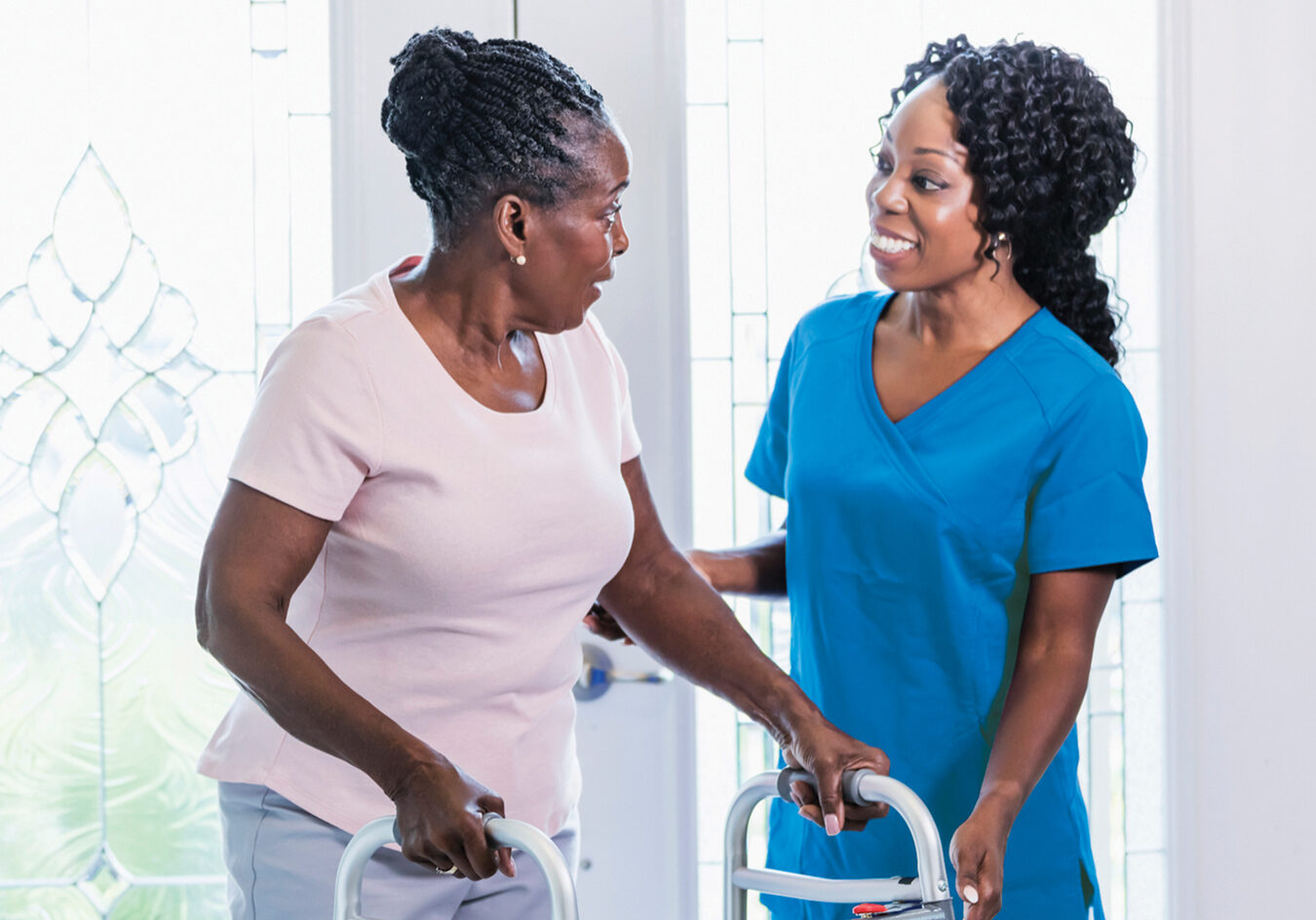 A healthcare worker visiting a senior African-American woman at home, helping her use a mobility walker. The patient is in her 70s and the nurse is a mature woman in her 40s.