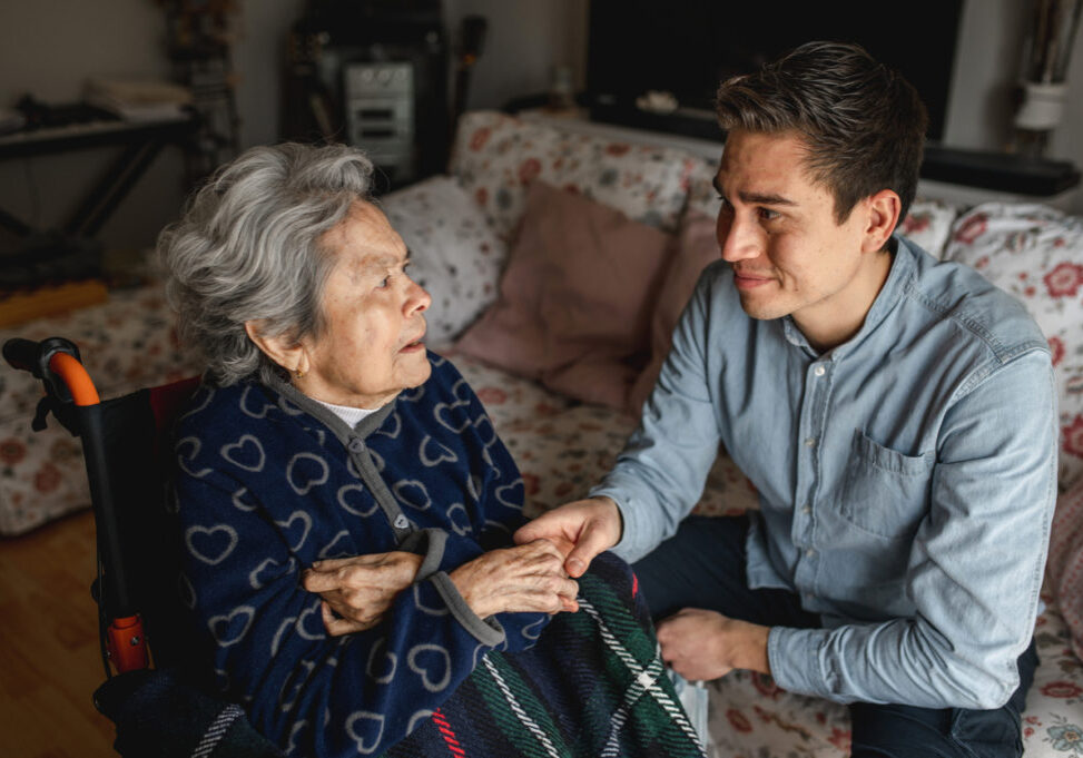Young man sitting next to an old sick aged woman in wheelchair taking her hands while talking and smiling. Family, home care concept.