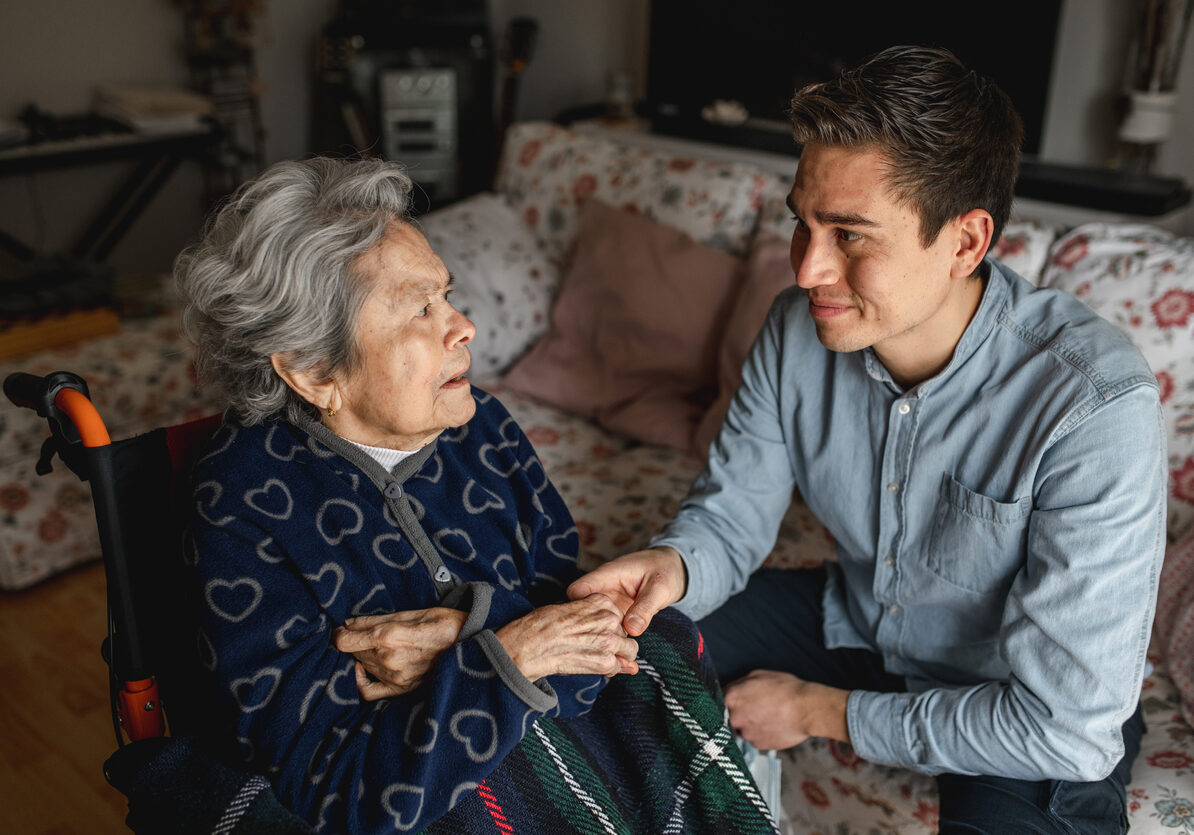 Young man sitting next to an old sick aged woman in wheelchair taking her hands while talking and smiling. Family, home care concept.