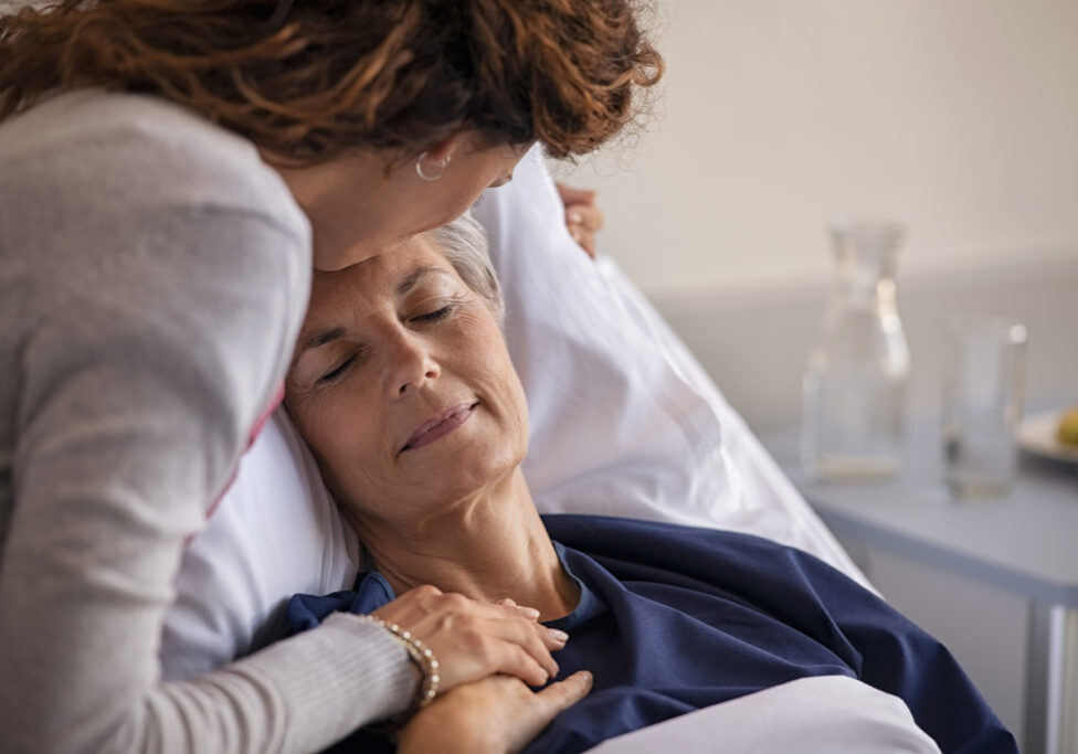 Close up of lovely daughter visiting and kissing sick mother on forehead while resting in hospital bed. Senior patient laying on gurney with eyes closed while daughter assuring a speedy recovery. Old mom patient being kissed while recovering from disease in nursing home.