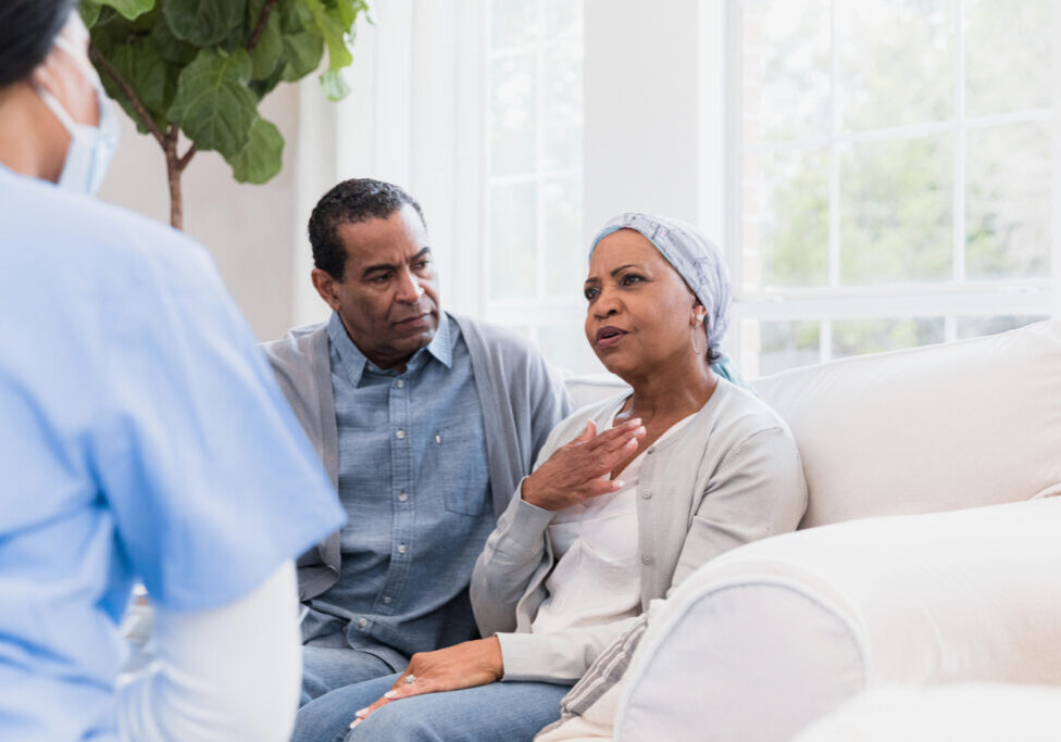 The senior adult husband watches worriedly as his wife describes her symptoms to the unrecognizable hospice nurse.