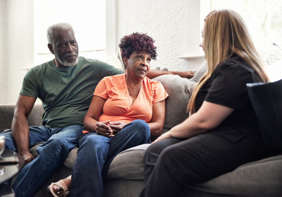 Caregiver talking with senior clients in family home Mid adult healthcare worker in uniform sitting on sofa with Black couple in casual attire and talking about services provided during house calls.