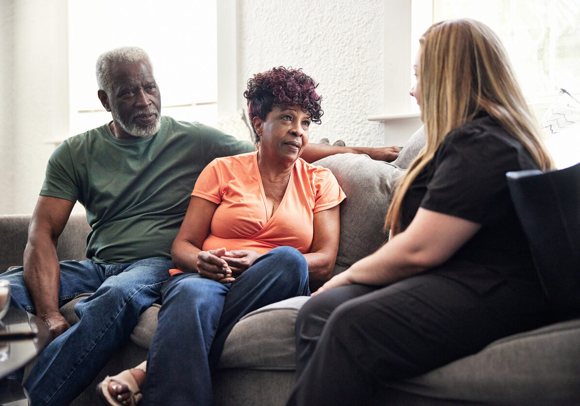 Mid adult healthcare worker in uniform sitting on sofa with Black couple in casual attire and talking about services provided during house calls.