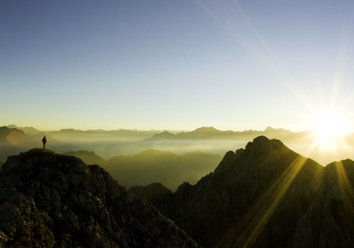 Photo of light rising over the mountains as a hiker looks on in the far distance; a representation of spirituality at end of life