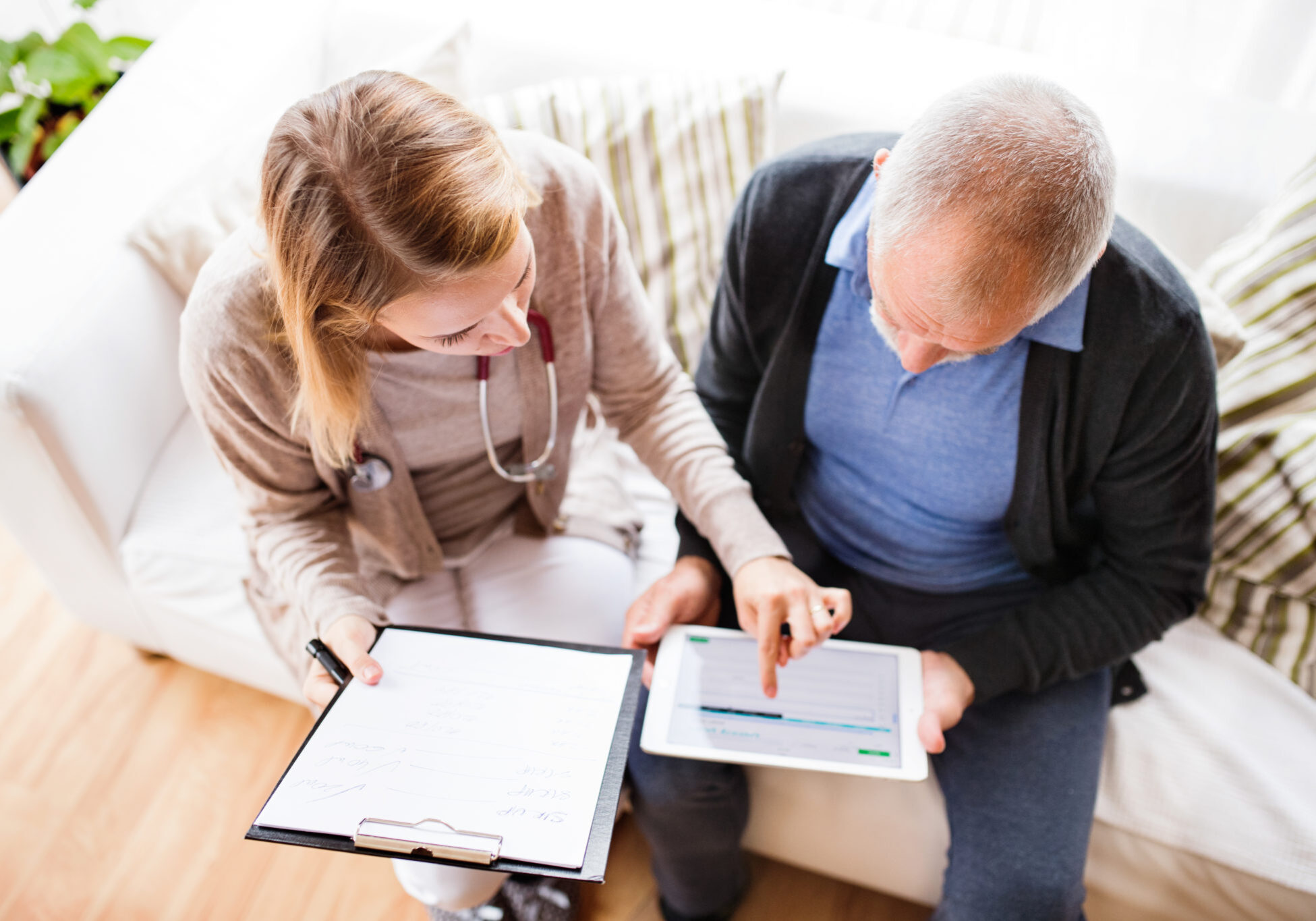 Health visitor and a senior man during home visit. A female nurse or a doctor showing test results on a tablet. High angle view.