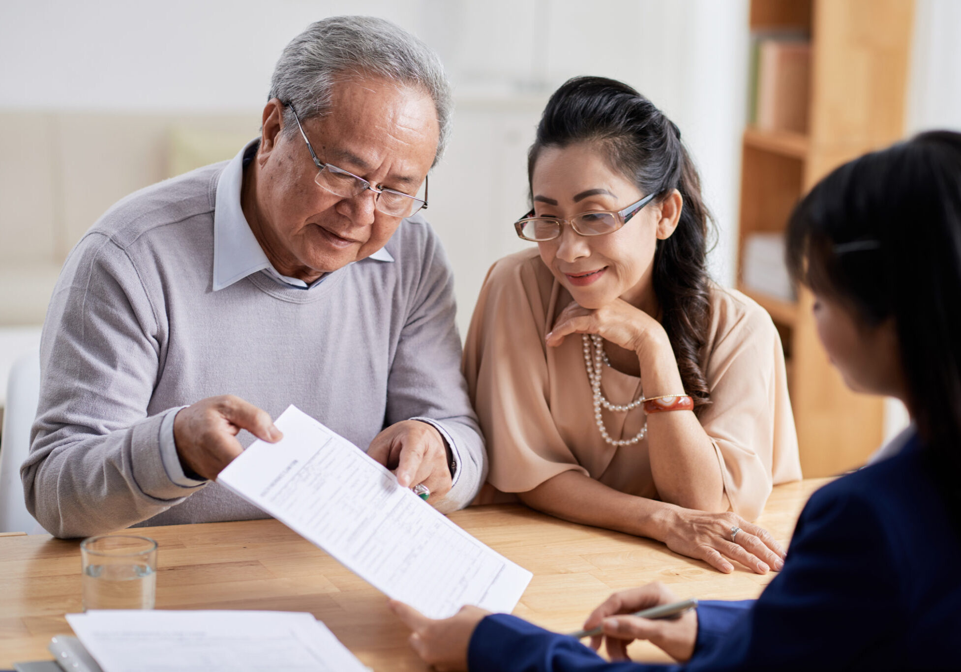 Concentrated senior man and his pretty wife studying terms of real estate purchase agreement while having meeting with realtor, interior of modern office on background