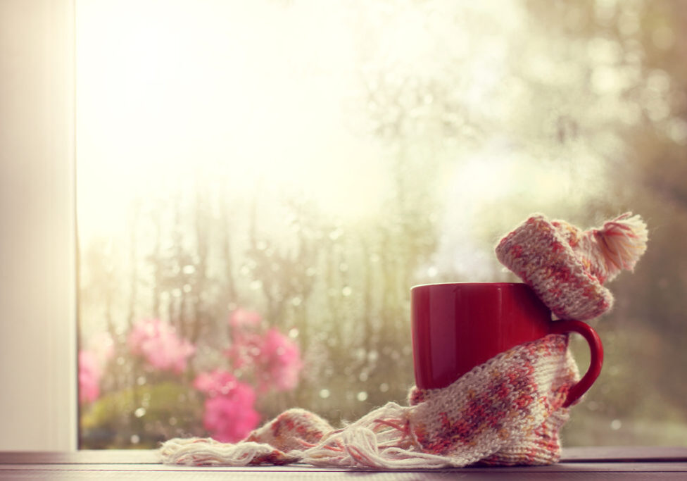 hot red mug in scarf and hat on the background window after the rain; symbolizing grief support during the holidays