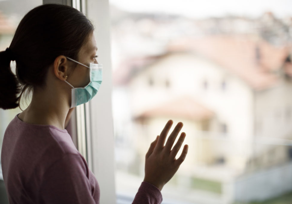 Portrait of sad teenage girl with face protective mask looking through the window at home Portrait of sad teenage girl with face protective mask looking through the window at home grieving during coronavirus COVID-19